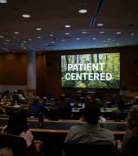 Audience in a dimly lit auditorium watches a large screen displaying "Patient Centered" over a serene forest backdrop, suggesting a focus on patient care.