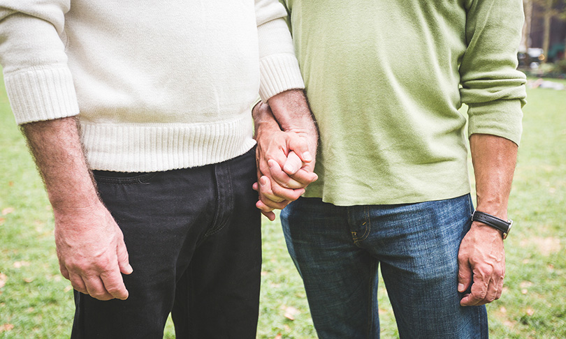 Two people standing outside, holding hands. One wears a white sweater and black pants, the other a green shirt and blue jeans. Their faces are not visible.