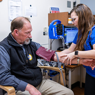 A healthcare worker checks a man's blood pressure in a clinical setting. The man sits calmly, and medical equipment is visible in the background.
