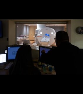 Silhouetted figures in a control room observe a medical imaging scanner through a window, highlighting technology and healthcare diagnostics.