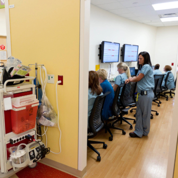 A nurse monitors five colleagues seated at desks facing wall-mounted screens in a hospital room. Medical equipment is visible in the foreground.