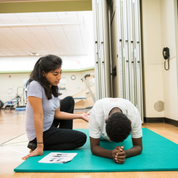 A woman guides a person doing a plank on a green mat in a gym. She gestures with her hand, suggesting instruction or encouragement. Exercise equipment is visible.