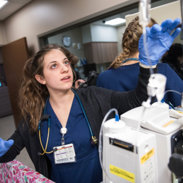 A nurse wearing blue scrubs and gloves adjusts an IV drip in a hospital setting. Her focused expression conveys a sense of care and professionalism.