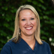 A woman with straight blonde hair smiles warmly in an outdoor setting. She wears a maroon top, with greenery and trees softly in the background.