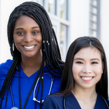 Two smiling healthcare professionals in blue scrubs stand outside. Both wear stethoscopes, conveying a sense of teamwork and approachability.