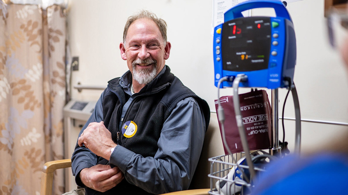 A smiling man in a medical setting sits beside a blue blood pressure monitor. He wears a black vest and appears relaxed, creating a positive and calm atmosphere.