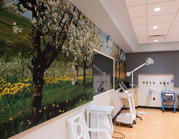 A medical room with wall art depicting a serene orchard. Equipment includes a tall examination light, a cart, and chairs, creating a calming atmosphere.