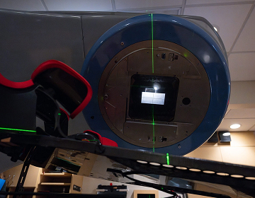 Close-up of a radiation therapy machine in a hospital room, displaying a metallic circular face with a central opening. Green alignment lasers are visible.