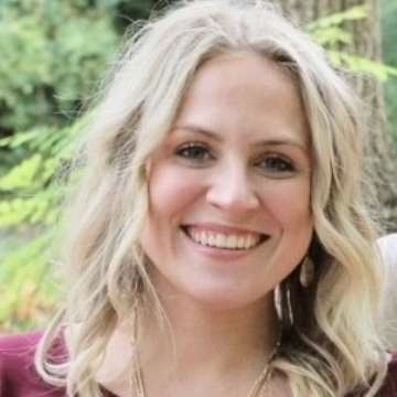 A woman with wavy blonde hair smiles warmly in an outdoor setting. She wears a maroon top, with greenery and trees softly blurred in the background.