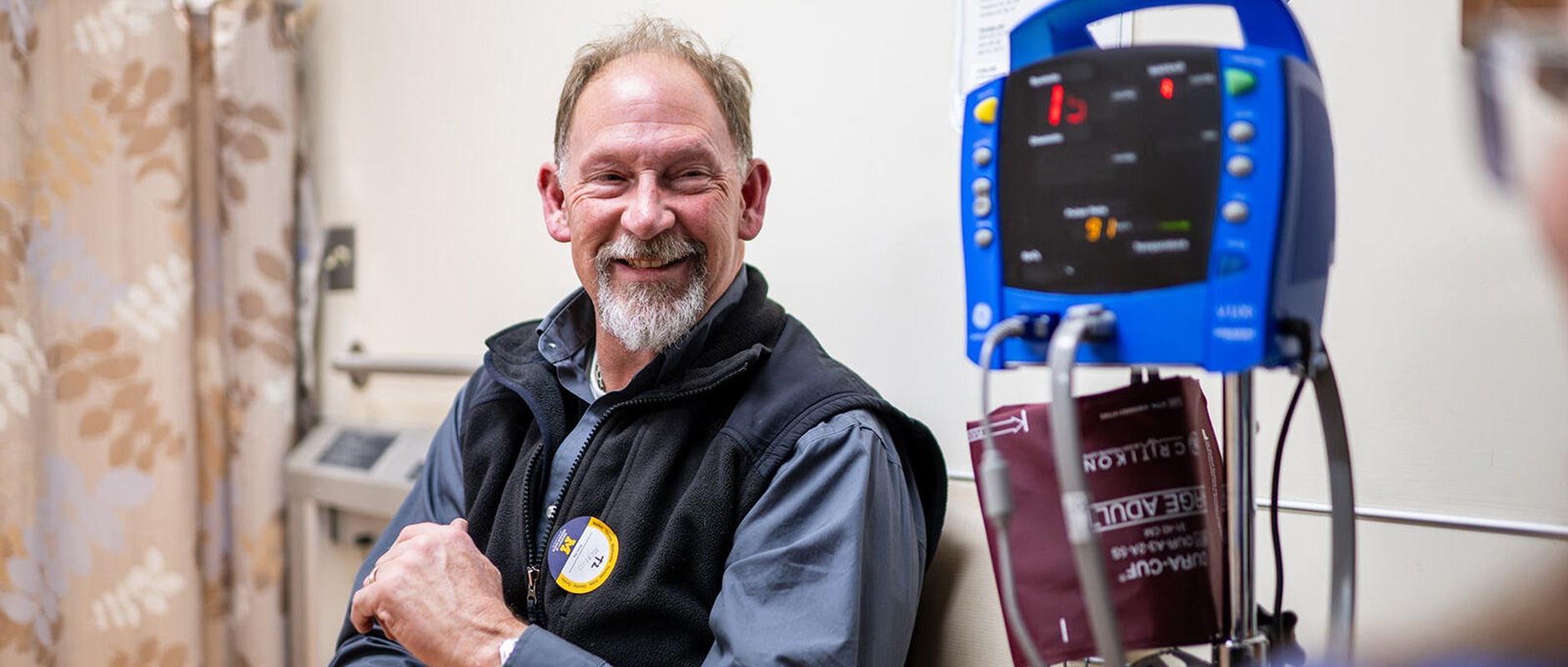 A smiling man in a medical setting sits beside a blue blood pressure monitor. He wears a black vest and appears relaxed, creating a positive and calm atmosphere.