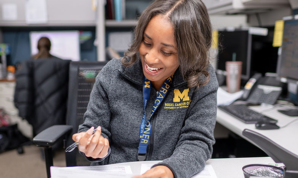 A smiling woman in an office, wearing a gray jacket with an "M" logo, holds a pen over papers. She exudes a positive, cheerful atmosphere.