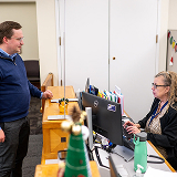 A man in a blue sweater talks to a woman seated at a desk with computers, files, and a green water bottle. Office setting, serious tone.