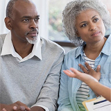 Elderly couple listens attentively to an advisor’s explanation, seated indoors. The woman gestures thoughtfully, creating an engaging and serious tone.
