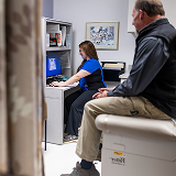 A patient sits on an exam table in a medical office, watching a nurse in blue scrubs typing on a computer. The room is tidy with a framed picture.