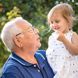 An elderly man with glasses joyfully holds a smiling young girl in a white dress. They share a tender moment outdoors with a green blurred background.