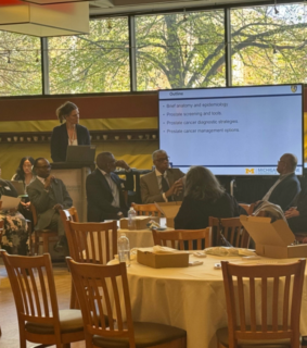A group of nine people sit in a conference room, engaged in a discussion. A presentation screen displays topics on prostate cancer. The atmosphere is professional.