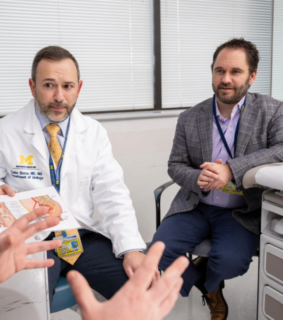 Two men sit in an office setting, one in a lab coat holding a medical diagram. They appear engaged in a professional discussion.