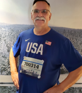 Man smiling in a blue "USA" shirt with bib number 700314, posing against a snowy forest mural. He exudes a sense of pride and enthusiasm.