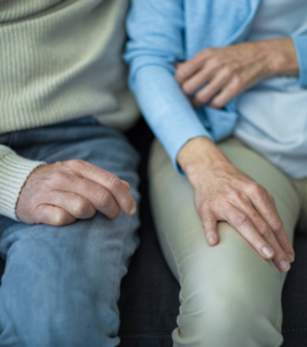 Elderly couple sitting closely on a couch, wearing casual clothes. Their hands rest gently on their own laps, conveying warmth and companionship.
