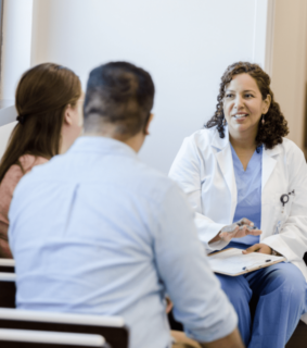 A doctor in a white coat and blue scrubs sits with a notepad, smiling and talking to a couple. The atmosphere is calm and supportive.