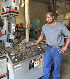 Man in gray shirt and blue jeans stands next to industrial machinery in a workshop. He smiles, suggesting pride in his work. Equipment and tools are visible.