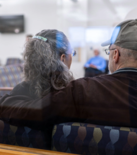 A couple sits closely, facing forward, with the man's arm around the woman. They are in a waiting room with soft lighting, conveying calmness and support.