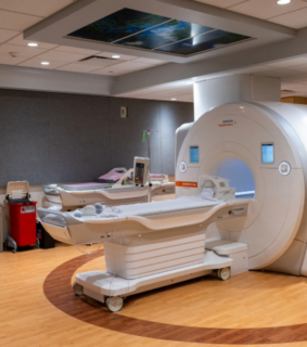 Modern MRI room with large scanner in the center, featuring soft lighting, wood-patterned flooring, and medical equipment, creating a clinical yet calming atmosphere.