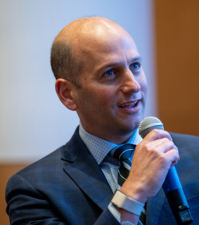 A man in a blue suit and tie speaks into a microphone at an indoor event, appearing engaged and professional, conveying an attentive atmosphere.
