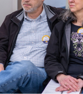 A couple sits closely on a patterned couch, wearing matching badges with numbers. They appear pensive and supportive, holding hands in a quiet moment.