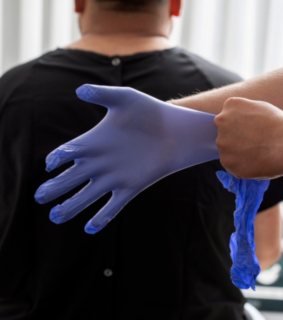 Person wearing blue gloves stands behind someone in a black shirt, suggesting a medical or professional environment. The tone is clinical and focused.