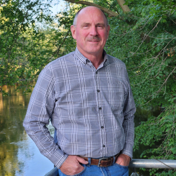 A man in a plaid shirt stands by a riverside railing, surrounded by lush green trees. He smiles warmly, creating a relaxed and natural atmosphere.