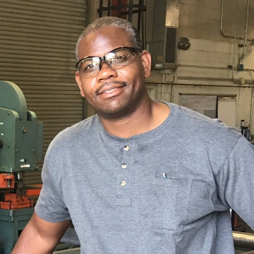 Man in gray shirt and blue jeans stands next to industrial machinery in a workshop. He smiles, suggesting pride in his work. Equipment and tools are visible.