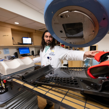 A healthcare professional in a white coat adjusts radiation therapy equipment at Michigan Medicine. A computer screen and various medical devices are visible, indicating a clinical setting.