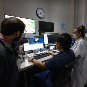 Three medical professionals in a radiation therapy setting are analyzing images on multiple computer screens. One points at the screen; all wear masks.