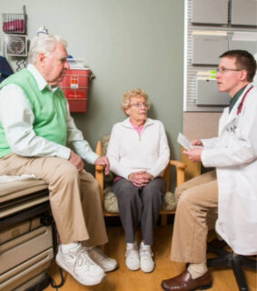 Doctor discusses with elderly couple in a medical office. The man sits on an exam table, while the woman sits on a chair, both attentive and serious.