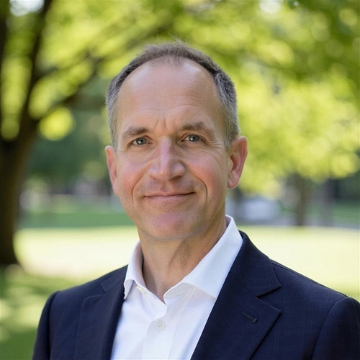 A smiling man in a white shirt and navy blazer with blurred green trees landscape in the background. He has short hair and is looking directly at the camera, conveying a professional and approachable demeanor.