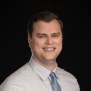 A smiling man in a light shirt and blue tie against a dark background. He has short hair and is looking directly at the camera, conveying a professional and approachable demeanor.