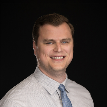 A smiling man in a light shirt and blue tie against a dark background. He has short hair and is looking directly at the camera, conveying a professional and approachable demeanor.