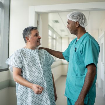 A doctor in teal scrubs and a surgical cap reassuringly touches the shoulder of a smiling man in a hospital gown in a bright corridor.