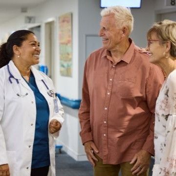 An elderly couple smiles warmly at a female doctor with a stethoscope, standing in a hallway. The atmosphere is positive and supportive.
