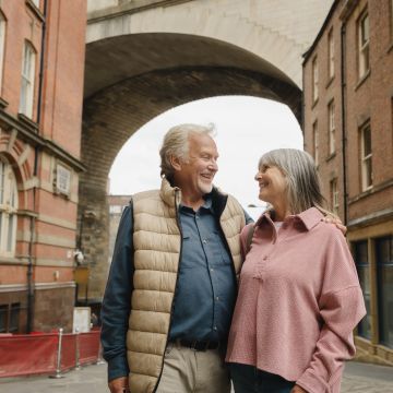 A smiling older couple stands arm in arm on a charming city street, under a stone bridge. The mood is warm and affectionate, suggesting joy and companionship.