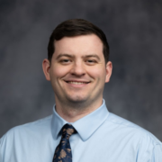 A man in a light blue shirt and patterned tie smiles warmly against a dark, blurred background, conveying a professional and approachable tone.