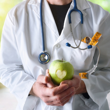 A doctor in a white coat holds a green apple with a heart shape carved into it. A stethoscope and measuring tape peek from the coat pocket.
