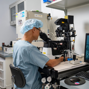 A person wearing surgical attire and a hair cap examines a sample using a microscope in a laboratory setting. The room contains medical equipment and a computer displaying data.