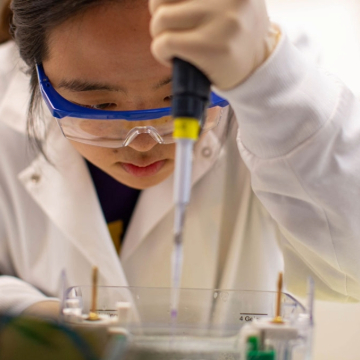 Researcher in a lab coat and safety goggles uses a pipette with focus and precision. The scene conveys concentration and scientific inquiry.