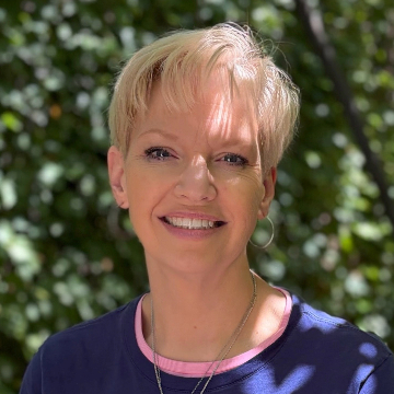 A woman with short blonde hair smiles warmly in an outdoor setting. She wears a maroon top, with greenery and trees in the background.